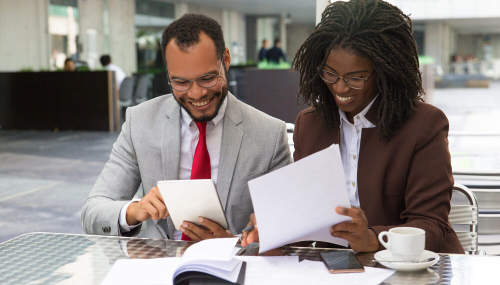 Cheerful business colleagues checking documents during coffee break. Business man and woman sitting in cafe, reading documents and using tablet. Paperwork concept